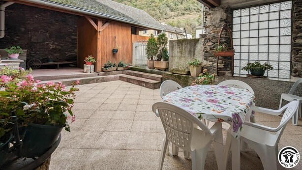 Une terrasse avec une table et des chaises, entourée de plantes en pot et d’un mur de pierre.