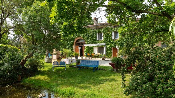 Ein üppiger Garten mit Teich, ein Essbereich mit Sitzgelegenheiten im Freien und ein Gebäude mit überdachter Terrasse.