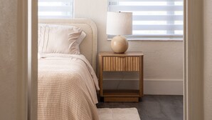 Ein Schlafzimmer mit einem Bett, einem Nachttisch, einer Lampe und zwei Fenstern mit Jalousien.