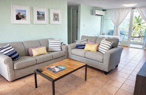 A living room with two grey sofas, a wooden coffee table, and a balcony with a view of palm trees.