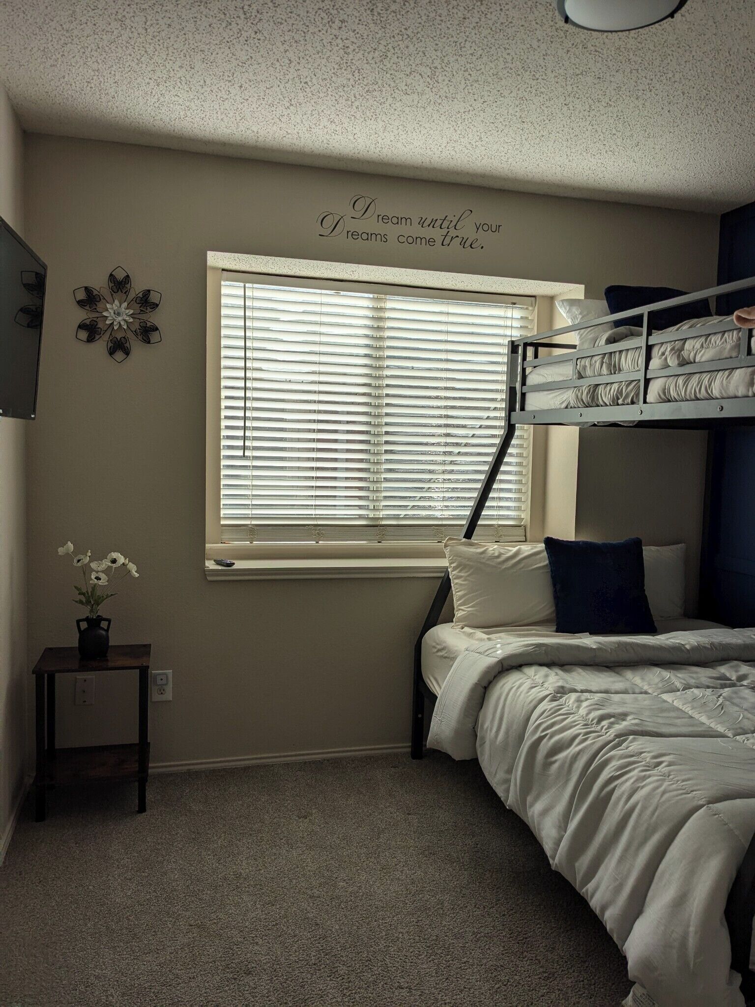 Um quarto com cama beliche, janela com persianas, uma mesinha com um vaso de flores e uma frase motivacional na parede.