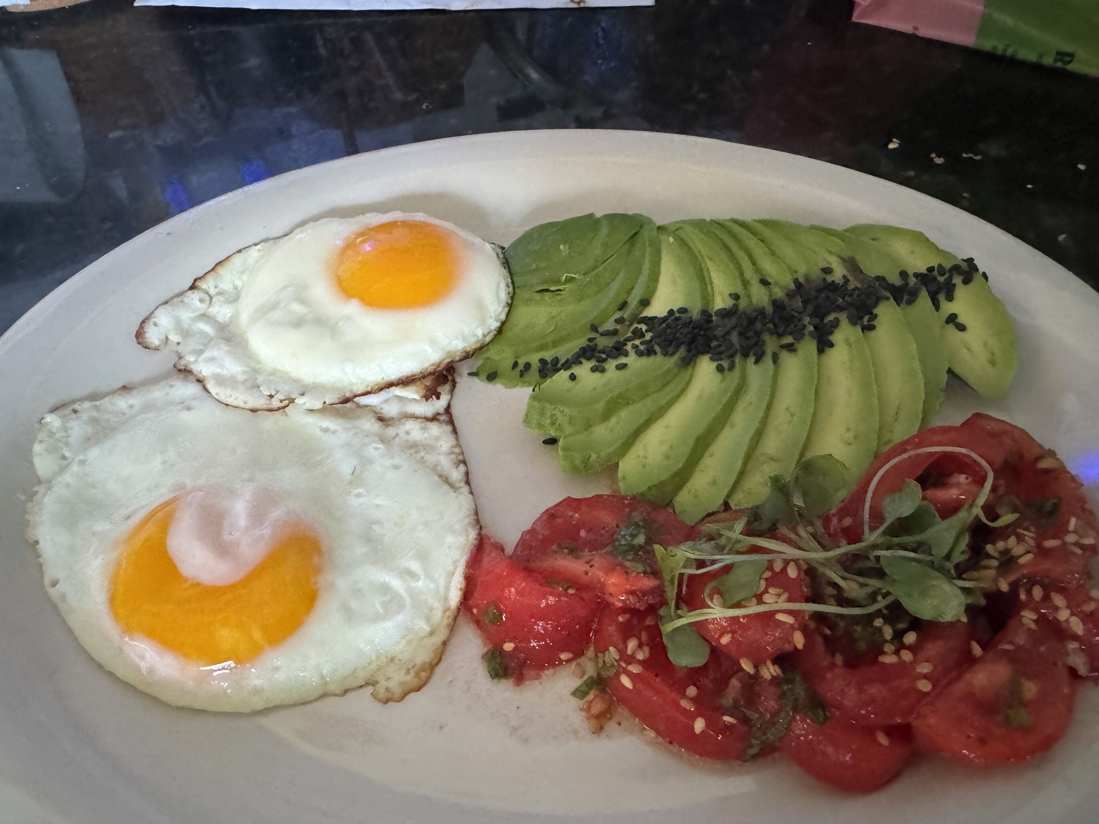 Un piatto con due uova all'occhio, avocado a fette guarnito con semi di sesamo nero, e un'insalata fresca di pomodori e verdure.