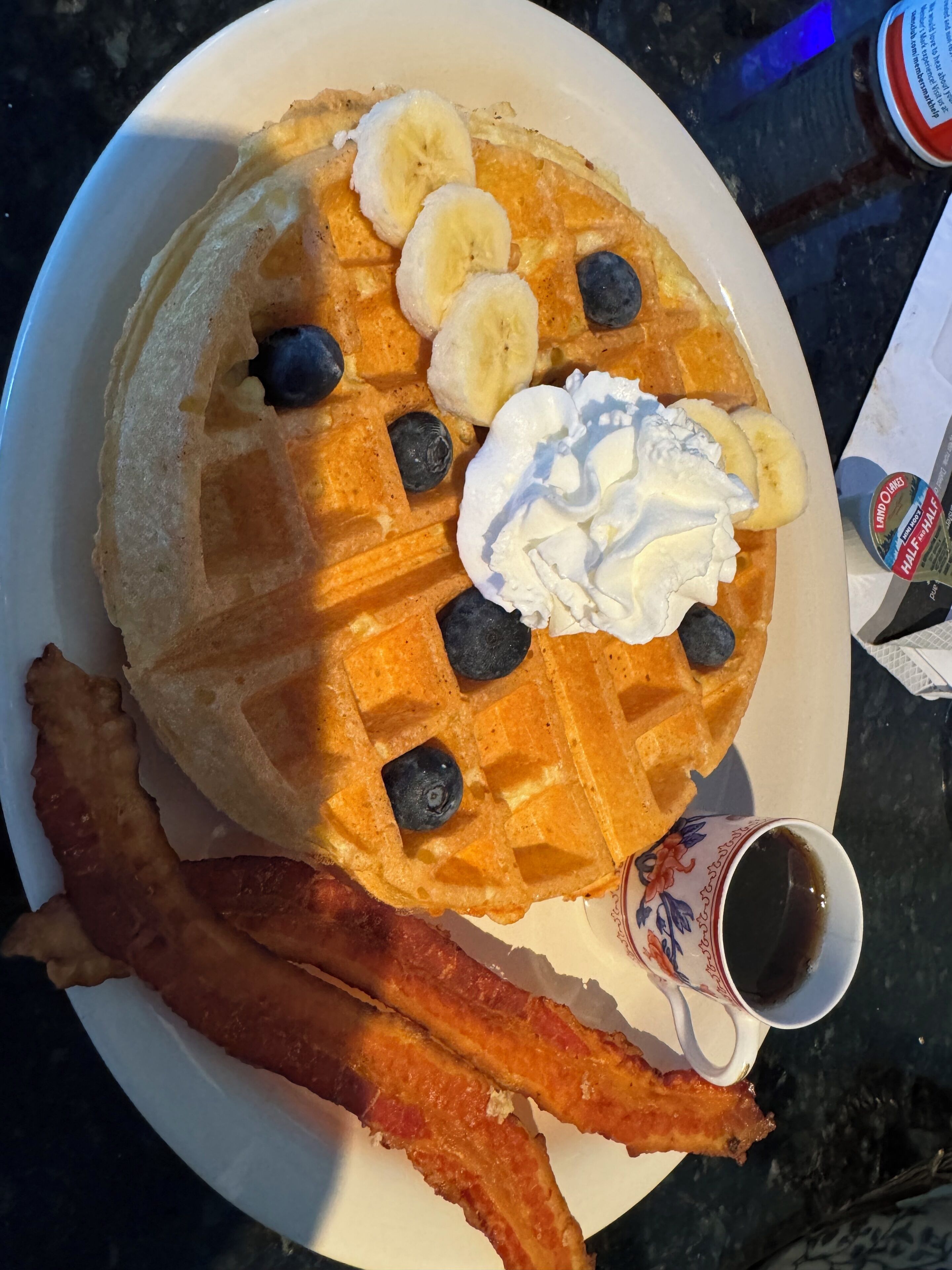 A plate with pancakes topped with banana slices, blueberries, whipped cream, and bacon strips.