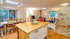 A modern kitchen with wooden cabinets, a dining table, and chairs. There is a large window providing natural light and a view of the outdoors.