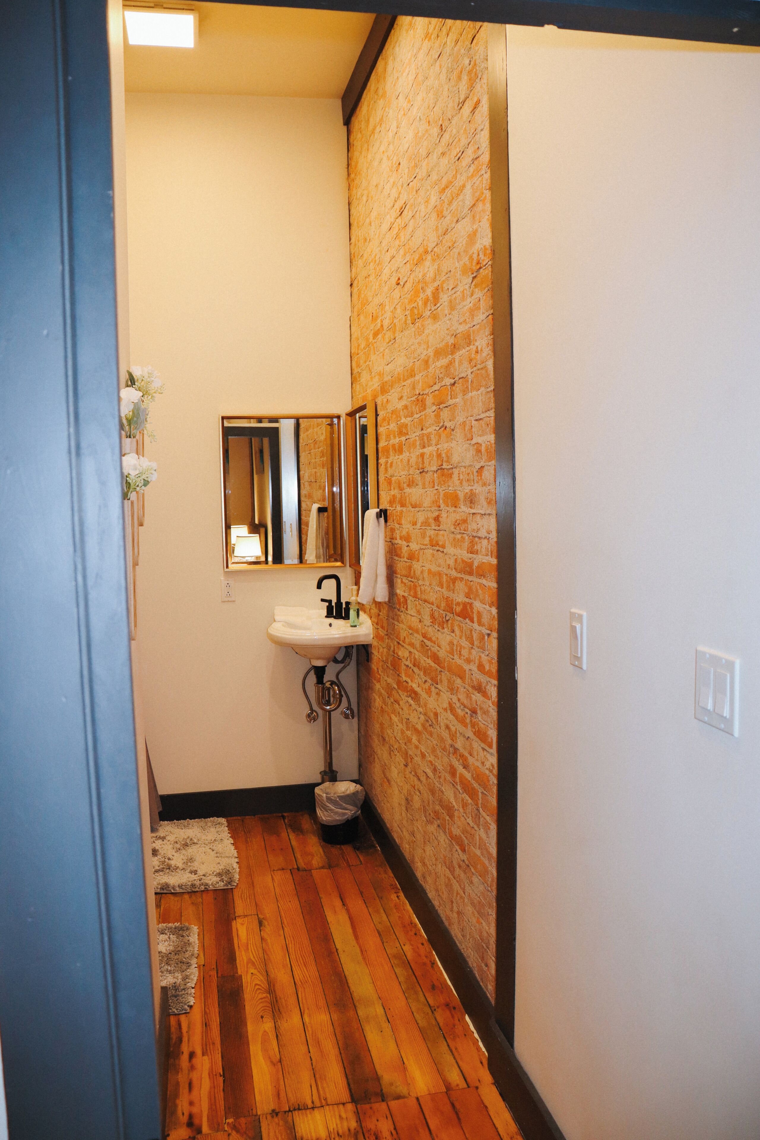 A bathroom with a wooden floor, a white sink, a mirror, and a brick wall.