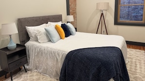 A bedroom with a bed, nightstands, a lamp, a painting, and a window with a brick wall view. - Boujee on Third: 2-bedroom condo in Beautiful Downtown Marion  (Marion)