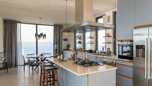 A modern kitchen with a central island, stainless steel appliances, and a large window offering a view. - Villa Peninsula, Luxury accommodation with breathtaking Sea and Mountain views  (Plaka)