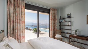 A bedroom with a bed, a desk, and a view of a pool and beach. - Villa Peninsula, Luxury accommodation with breathtaking Sea and Mountain views  (Plaka)