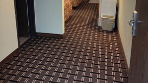 Et hotelmøbleret gangmiljø med et mønstret tæppe, seng med et mønsterbetrukket sengetæppe, en stol, en skraldespand og en dør med lampe.