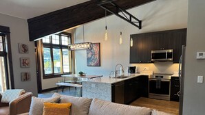 A modern living room with a beige sofa, a dining area, and a kitchen with dark cabinetry and stainless steel appliances. - Amazing Views - Close to it All! (Big Sky)