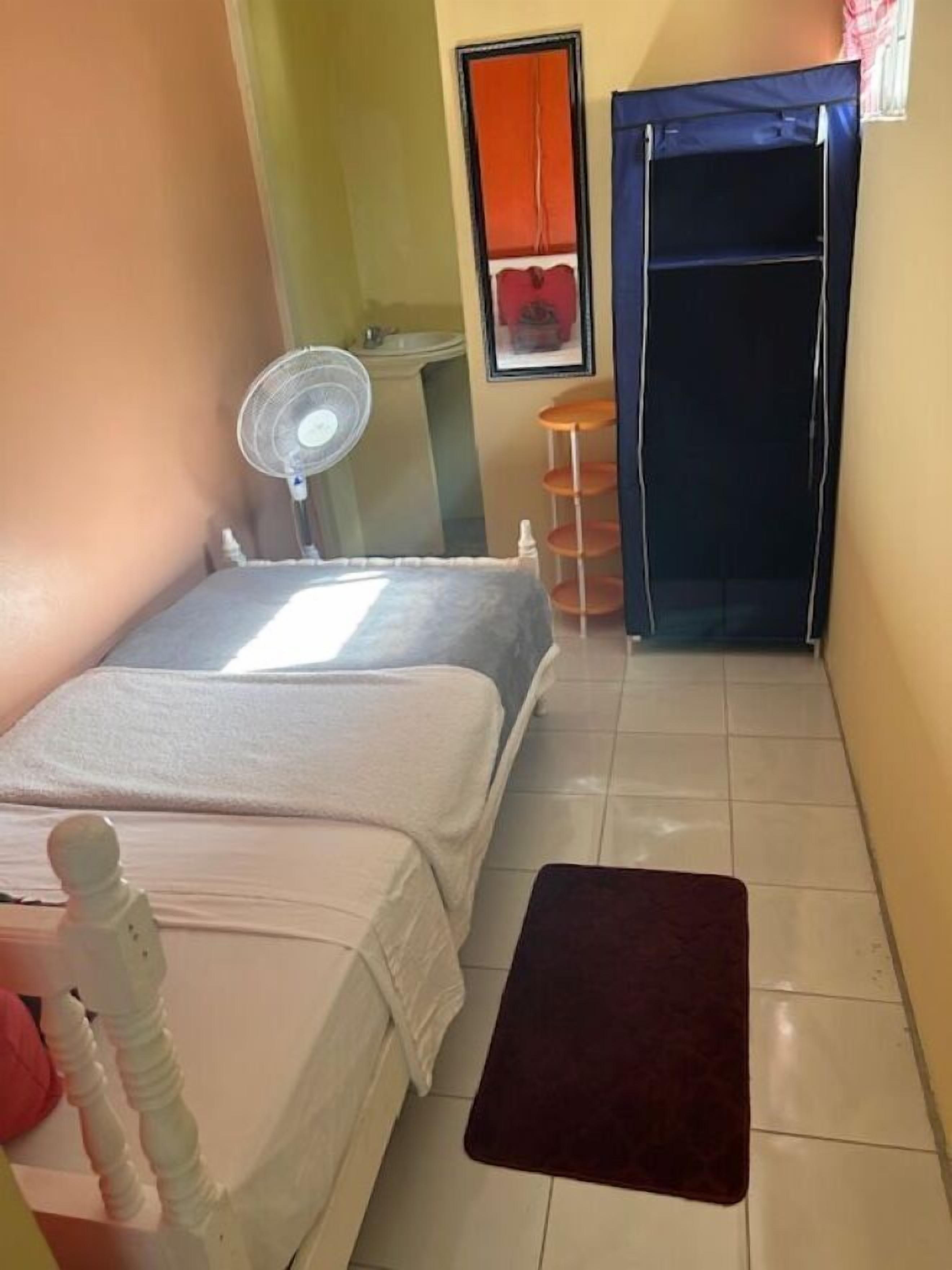 A small, well-lit room with two beds, a standing fan, a small table, and a wardrobe.