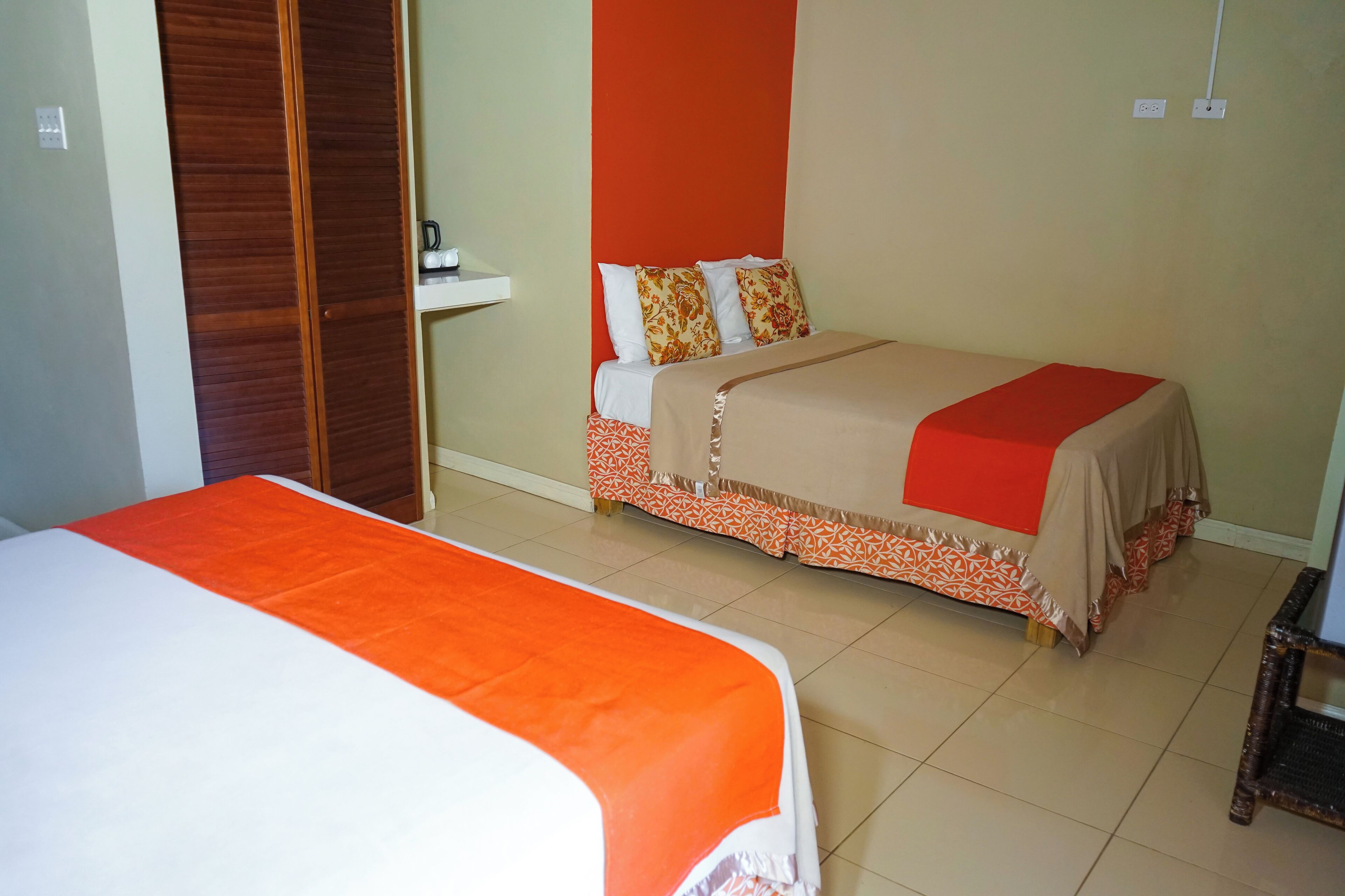 Een hotelkamer met twee bedden, waarvan er een een oranje sprei heeft en de andere een beige. Er is een houten garderobe en een klein tafeltje met een lamp.