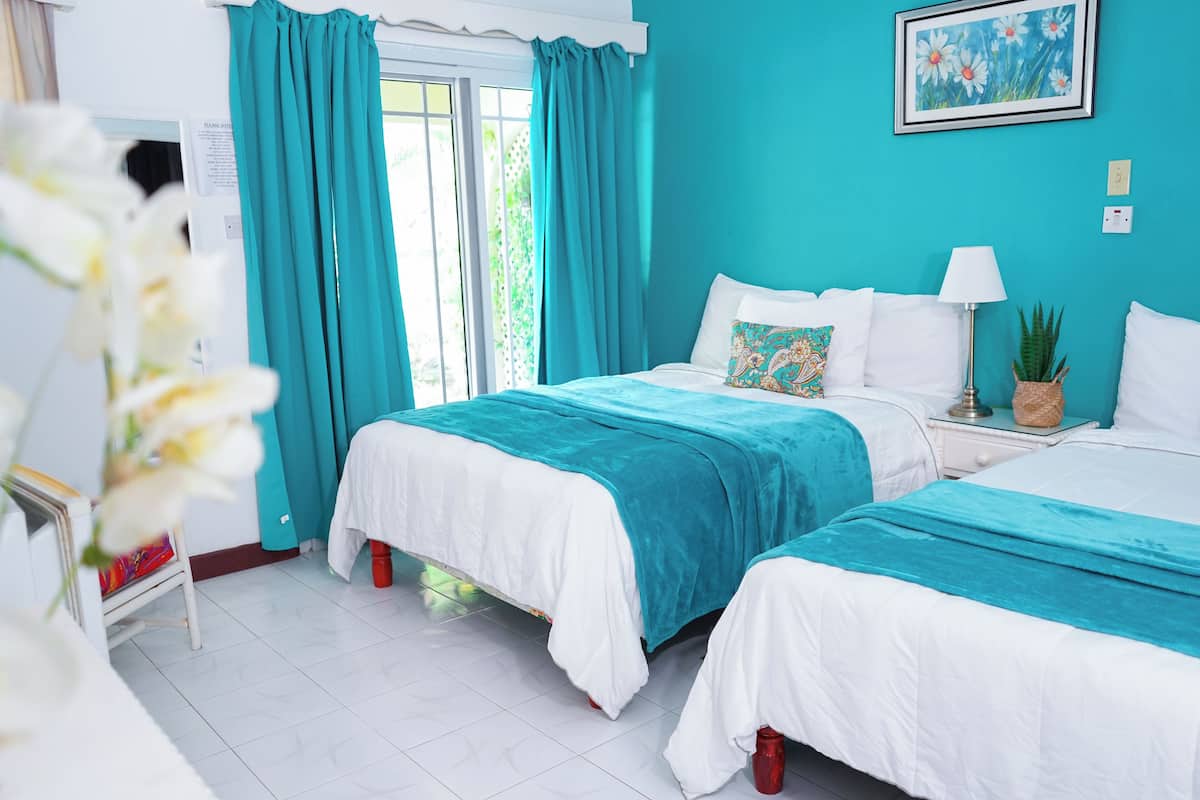Un dormitorio con dos camas, cortinas color turquesa, una lámpara blanca y un cuadro con flores en la pared.