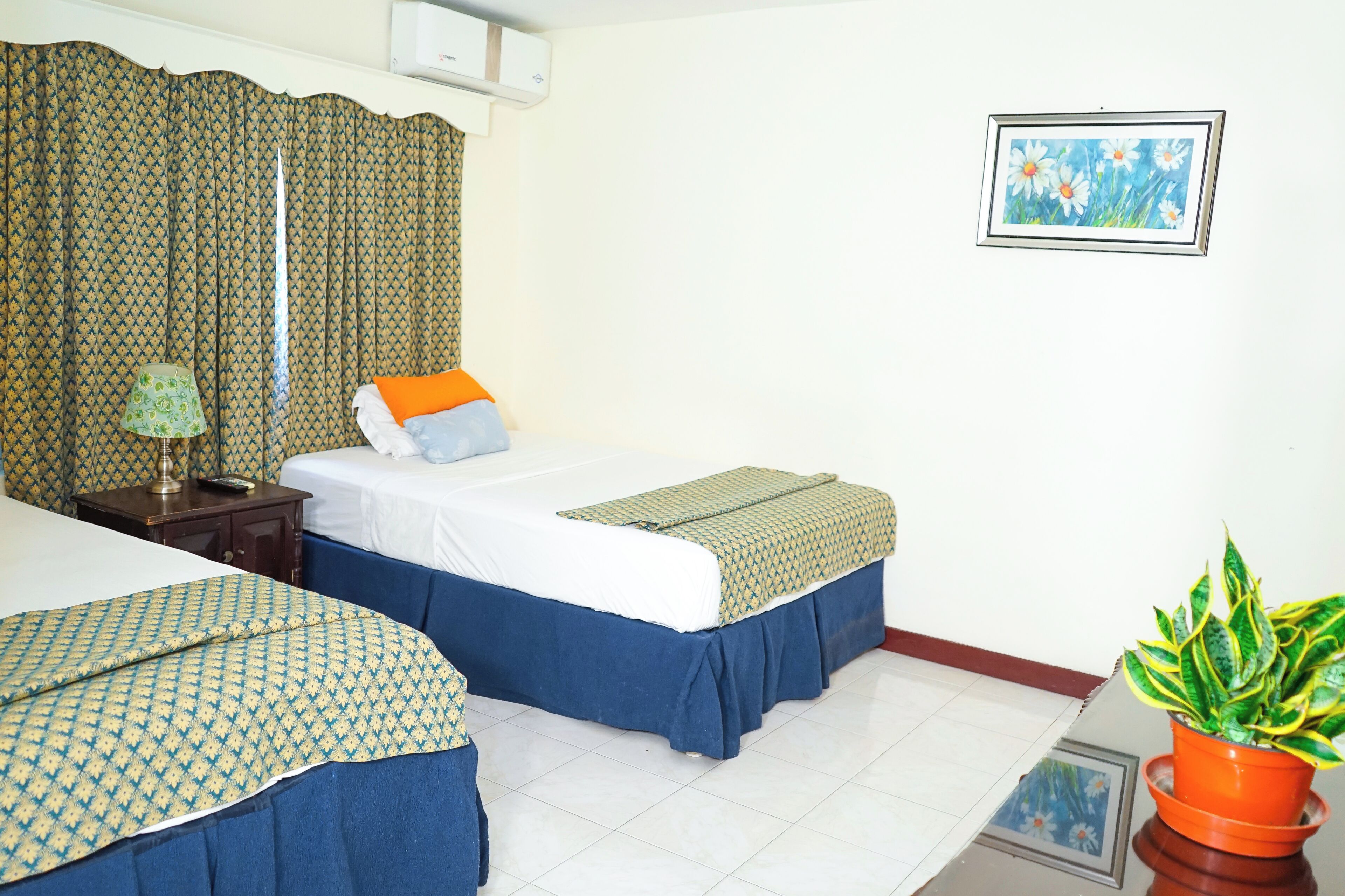 A hotel room with two beds, a nightstand, a lamp, a potted plant, and a framed picture on the wall.