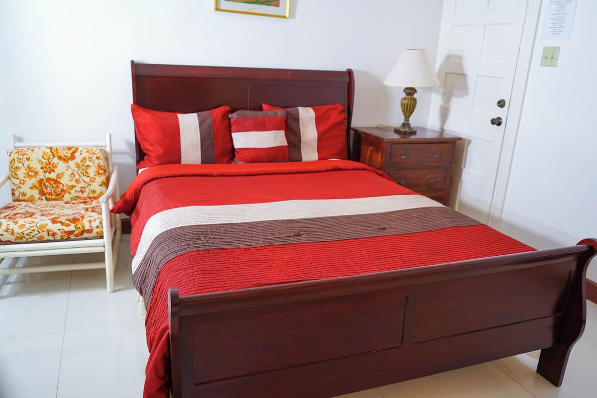 Una cama bien hecha con cabecera de madera, una mesita de noche con lámpara y una silla con cojín estampado.