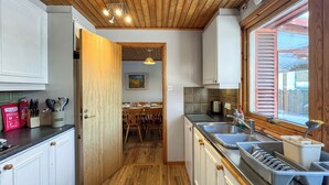 A compact kitchen with wooden flooring, white cabinets, a dining area with a table and chairs, and a window with wooden shutters.