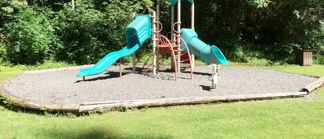 A playground with two slides and a climbing structure, surrounded by a grassy area and trees.