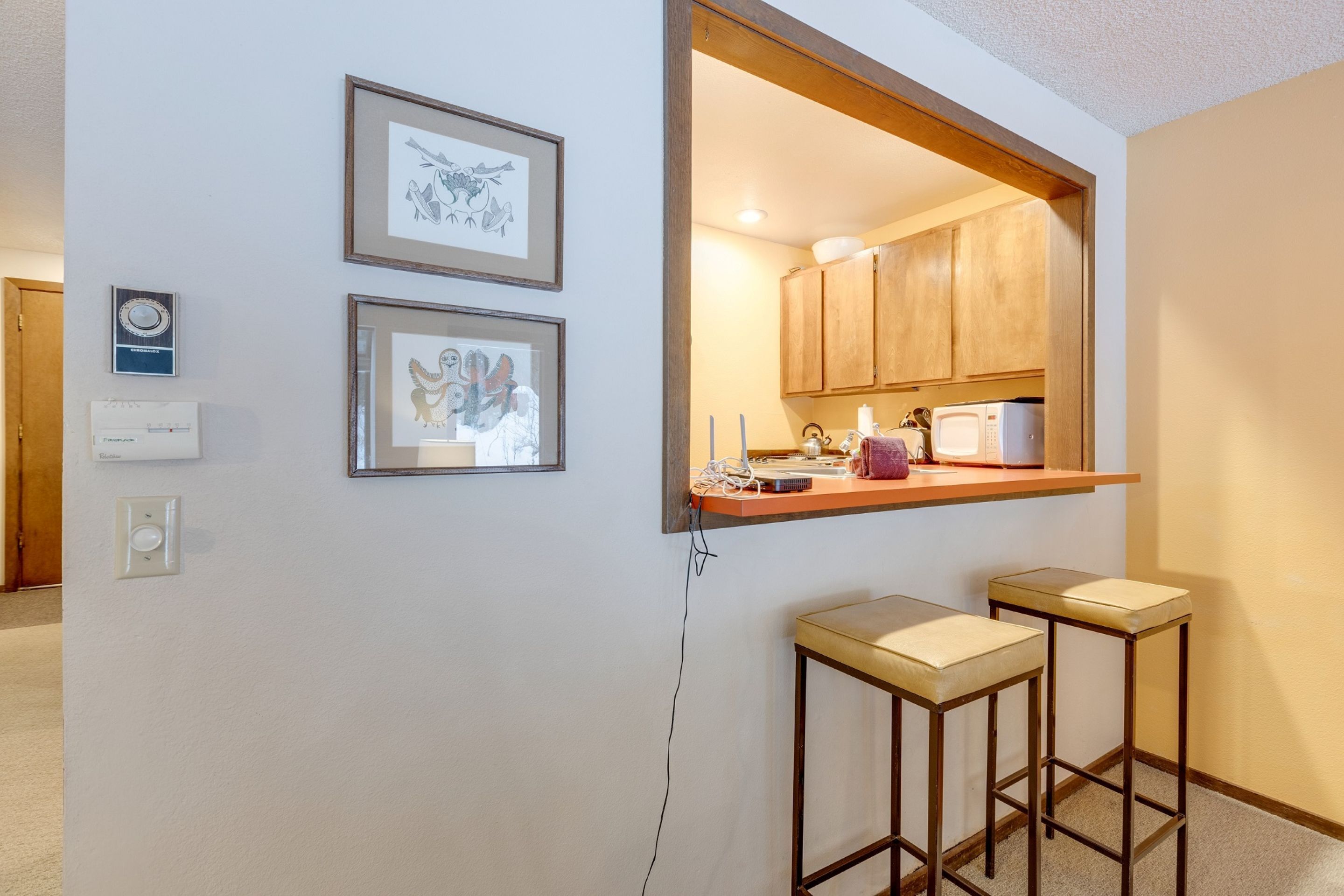 A kitchen with wooden cabinets, a countertop, and two bar stools. There are framed pictures on the wall and a thermostat on the wall.