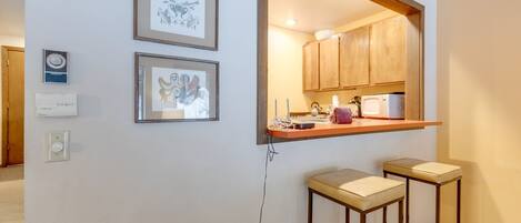 A kitchen with wooden cabinets, a countertop, and two bar stools. There are framed pictures on the wall and a thermostat on the wall.