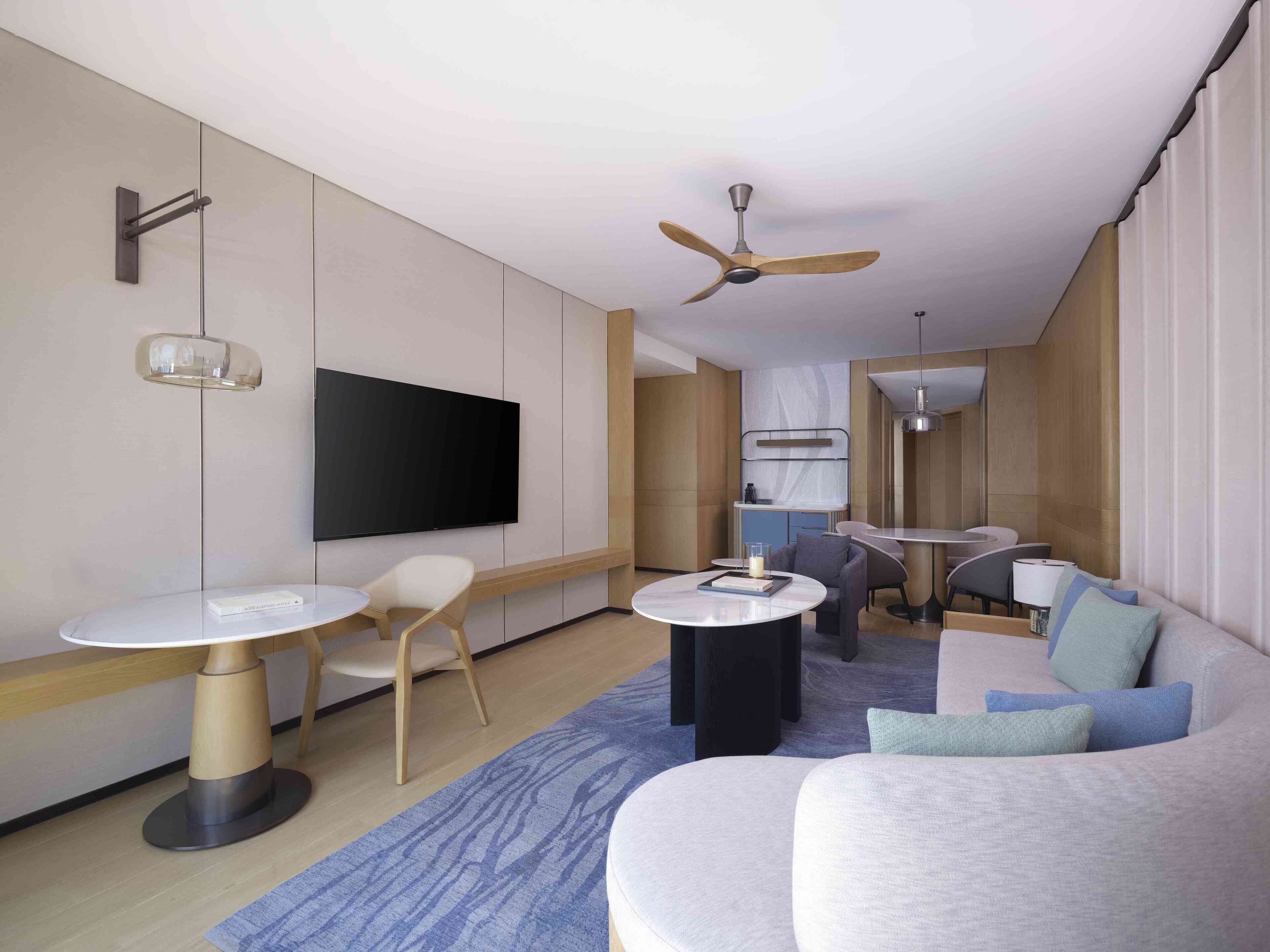 A modern hotel room with a flat-screen TV, a ceiling fan, a dining area with a table and chairs, and a sofa.