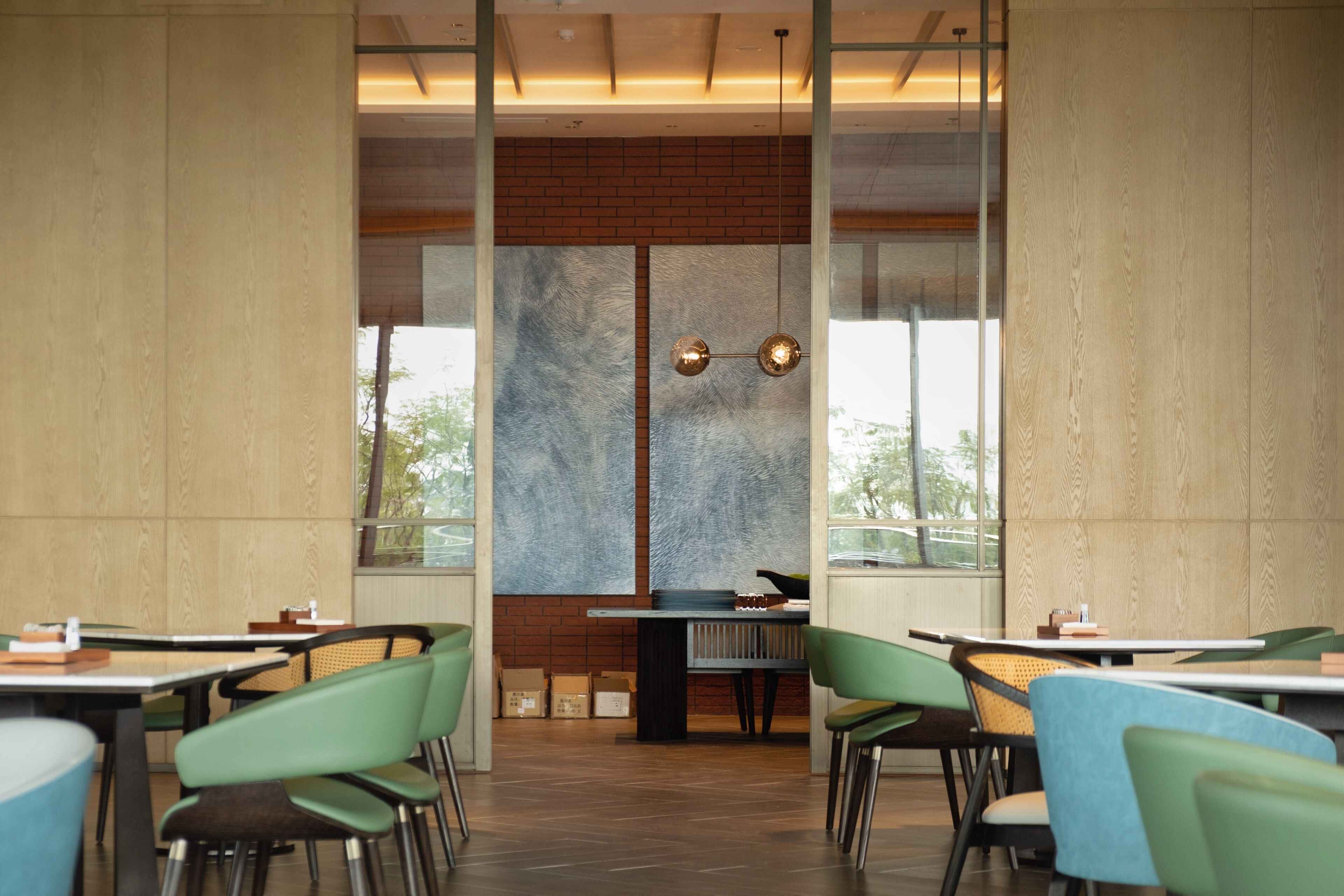 A modern restaurant interior with high ceilings, large windows, and a central brick wall. Tables and chairs are arranged for dining.