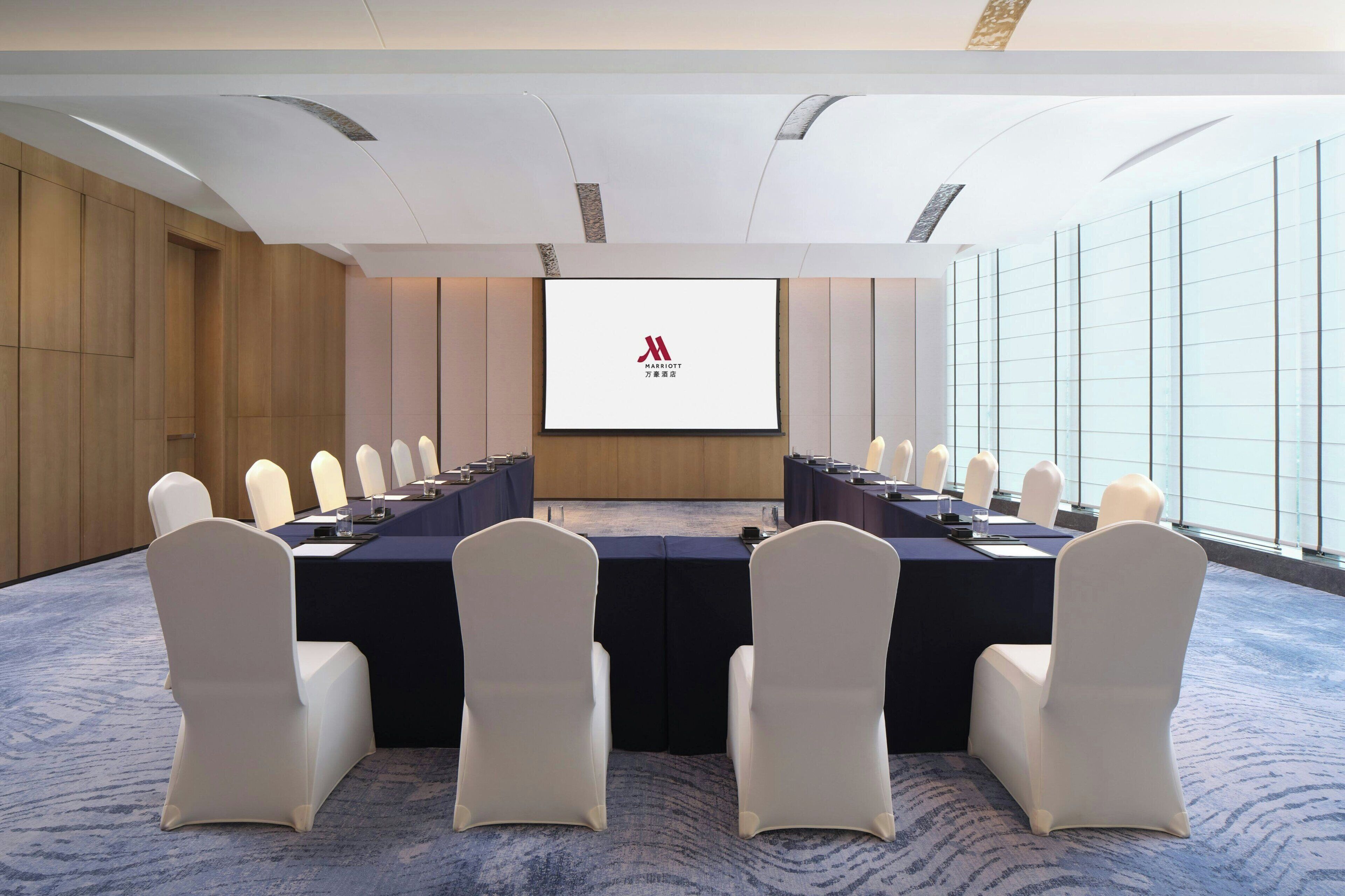 Un salón de conferencias con una larga mesa, sillas blancas y una pantalla para presentaciones.