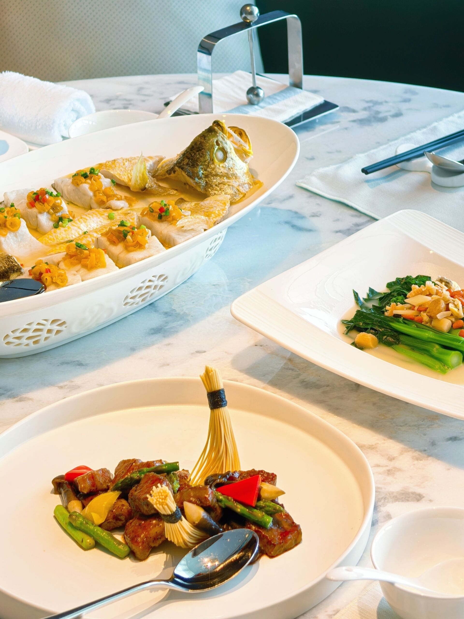 A dining table with three dishes: a fish dish, a vegetable stir-fry, and a pasta dish.