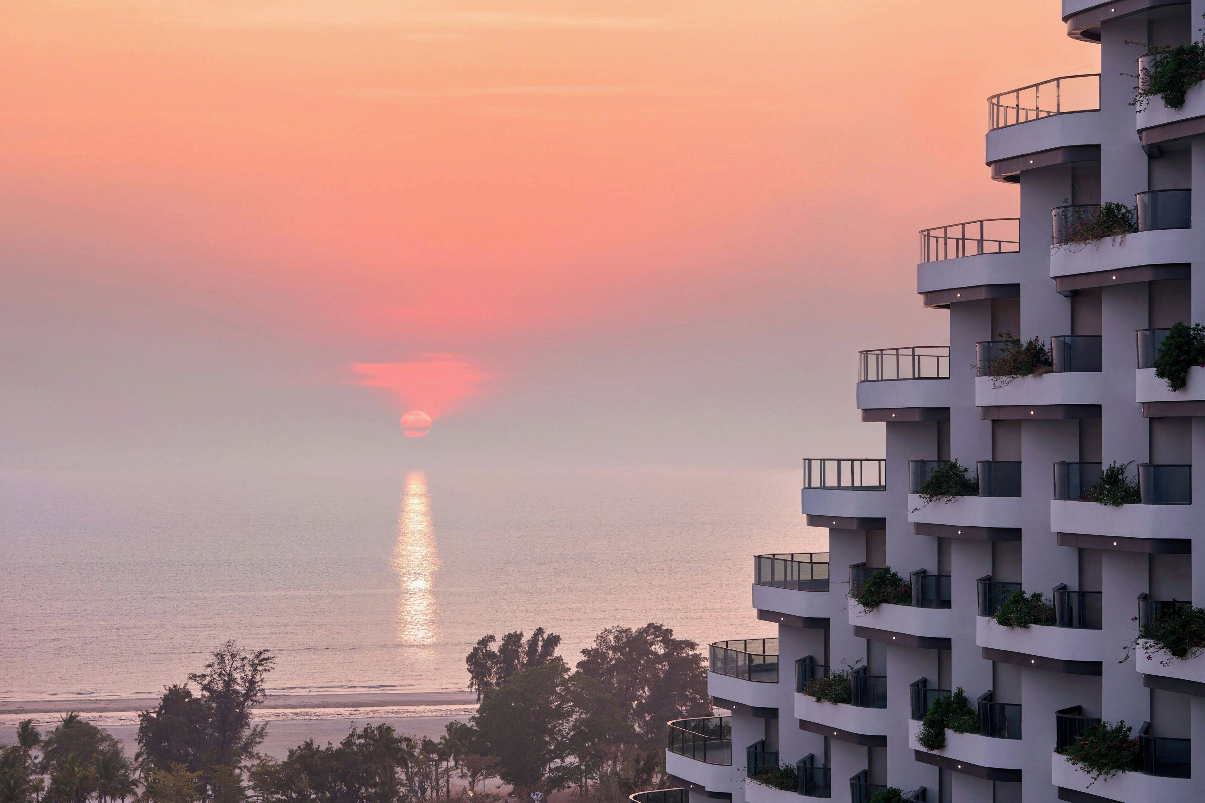 A modern building with multiple balconies, some with plants, overlooking a beach at sunset.