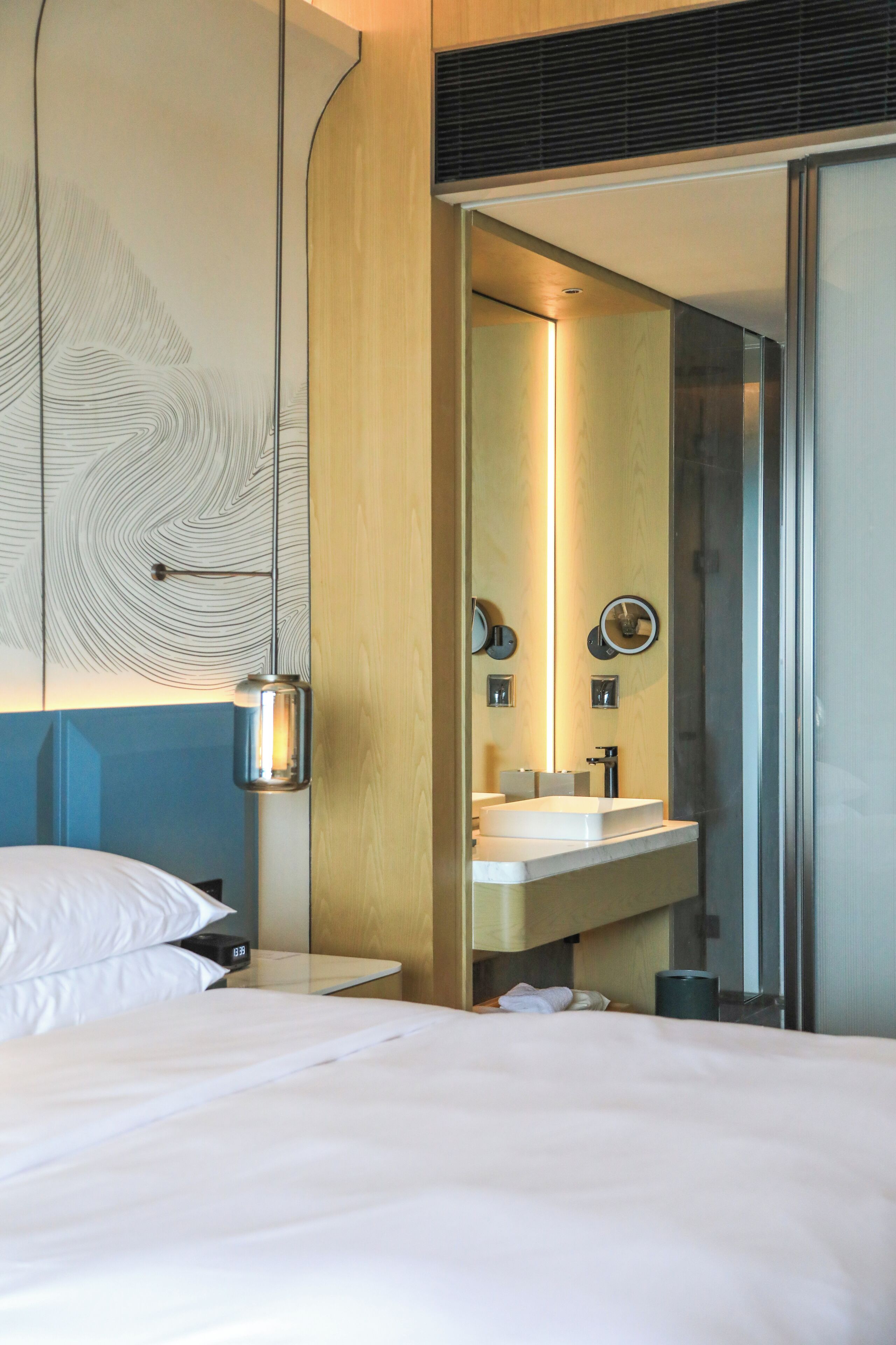 A modern hotel room with a large bed, a bedside table, a mirror, and a bathroom with a sink and shower.