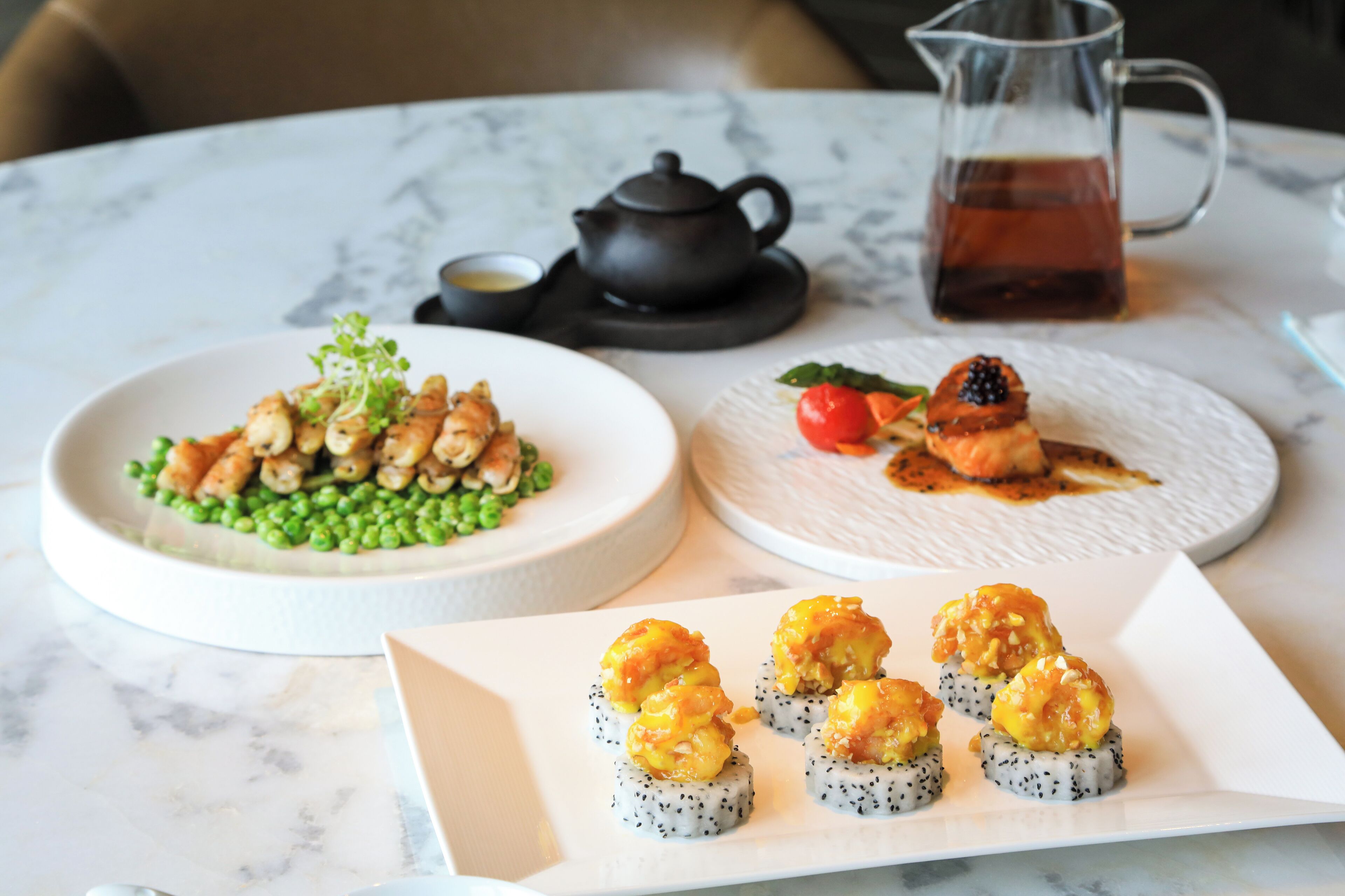 Una mesa con tres platos: uno de guisantes verdes y pollo, otro de cereza y salmón, y un plato de rollos de sushi coronados de camarones.