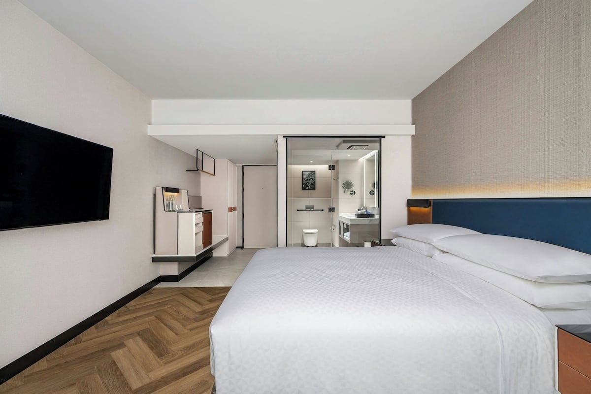 Una habitación de hotel moderna con una cama grande, un televisor de pantalla plana en la pared, piso de madera y un baño visible a través de una puerta abierta.