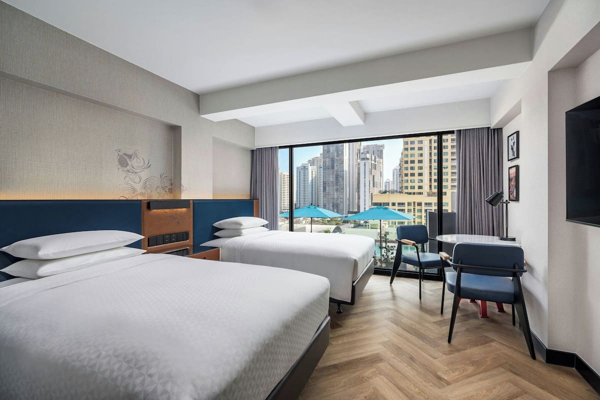 Habitación de hotel con dos camas, un escritorio, sillas y un ventanal con vistas a la ciudad.