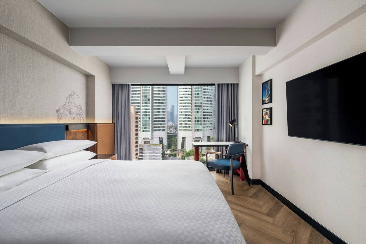 Habitación de hotel con una cama grande, un escritorio, una silla, un televisor y vistas de edificios altos.