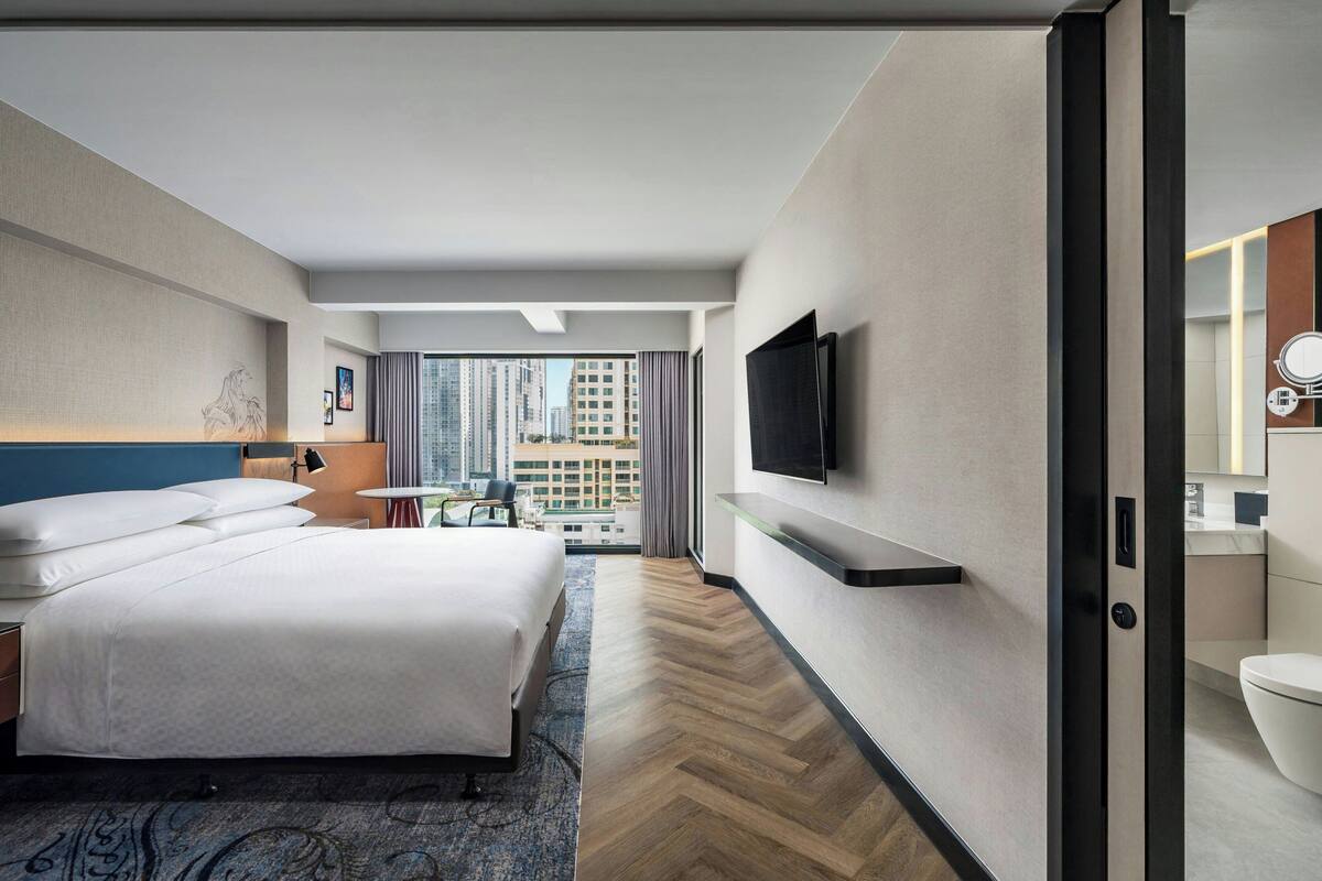 Una habitación de hotel moderna con una cama grande, un televisor de pantalla plana en la pared, una mesita con una silla y vistas a la ciudad a través de amplios ventanales.