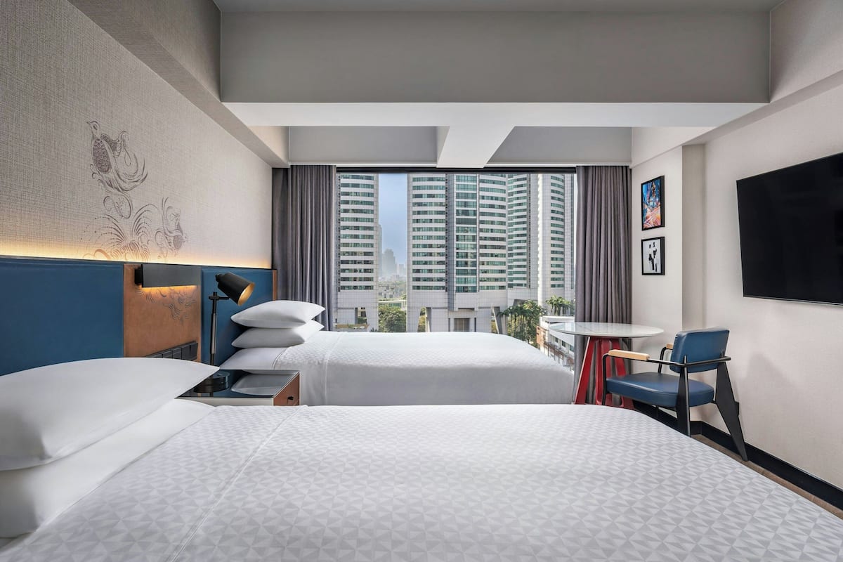 Habitación de hotel con dos camas, televisión, escritorio con silla y vista a edificios altos.