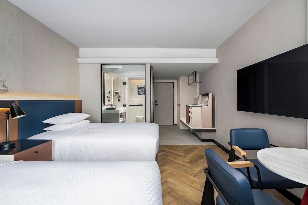 Habitación de hotel con dos camas, un escritorio, una silla, un televisor y un baño visible a través de una puerta abierta.