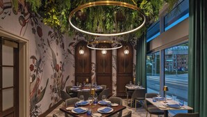 Un restaurante con una decoración moderna, que presenta un comedor con mesas preparadas para dos personas, un mural en la pared y vistas a la calle a través de grandes ventanales.