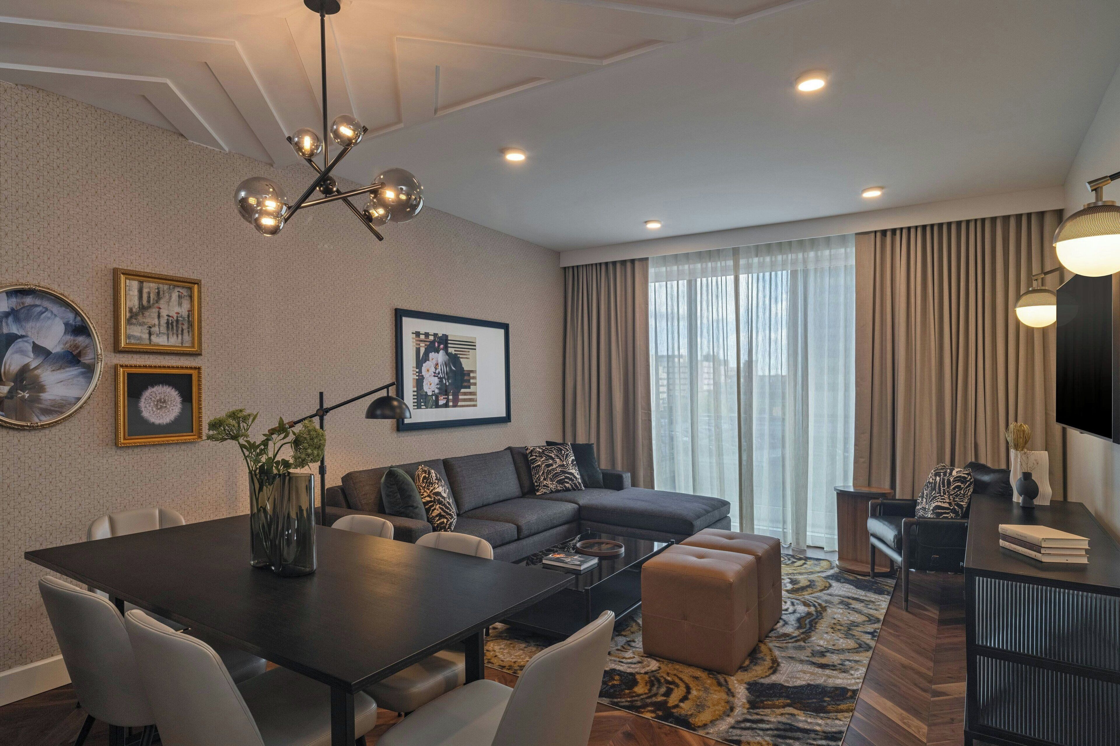 A modern living room with a sofa, coffee table, and dining table. There are framed pictures on the wall and a chandelier.