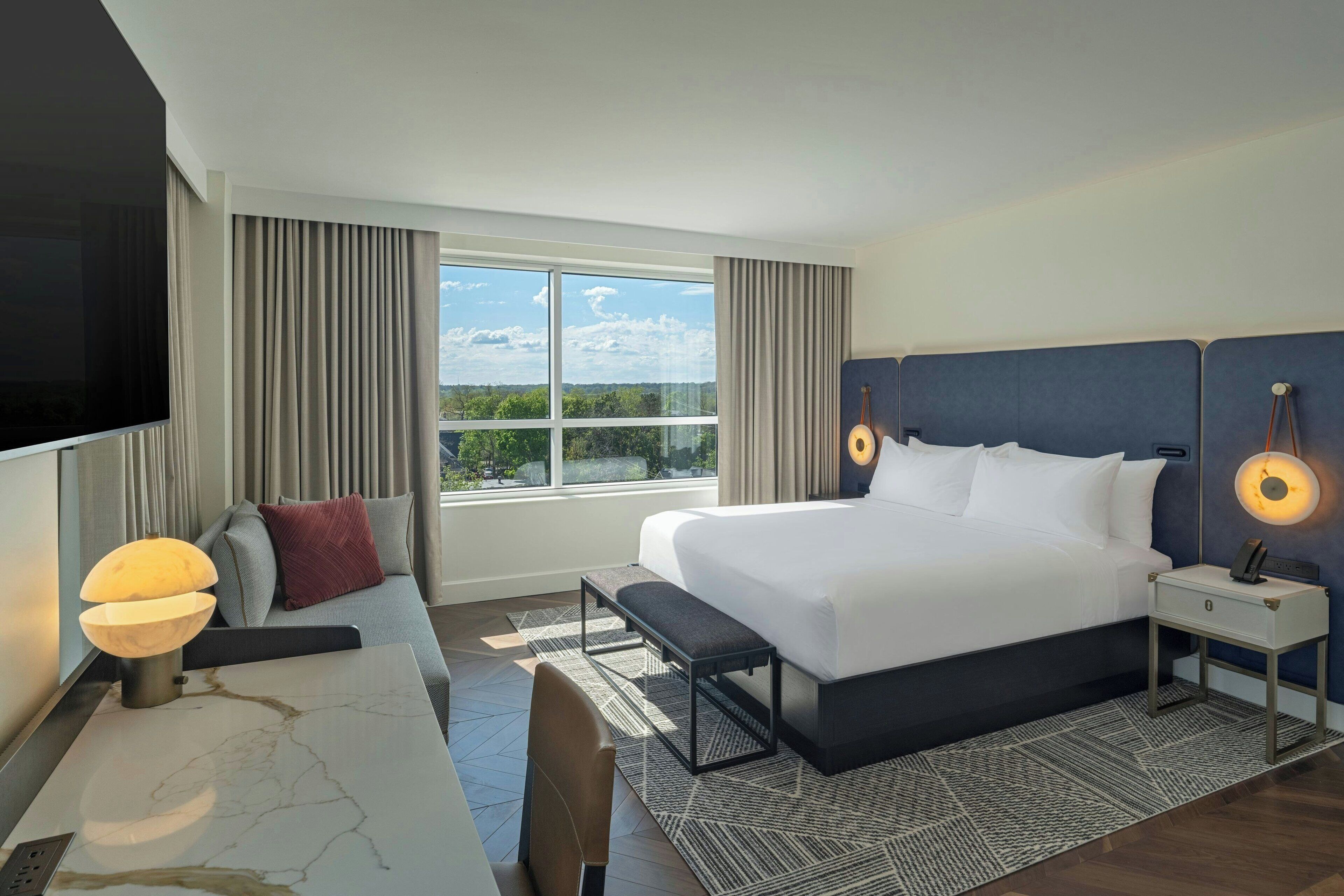 A hotel room with a large bed, a desk with a lamp, a TV, and a view of the outdoors.