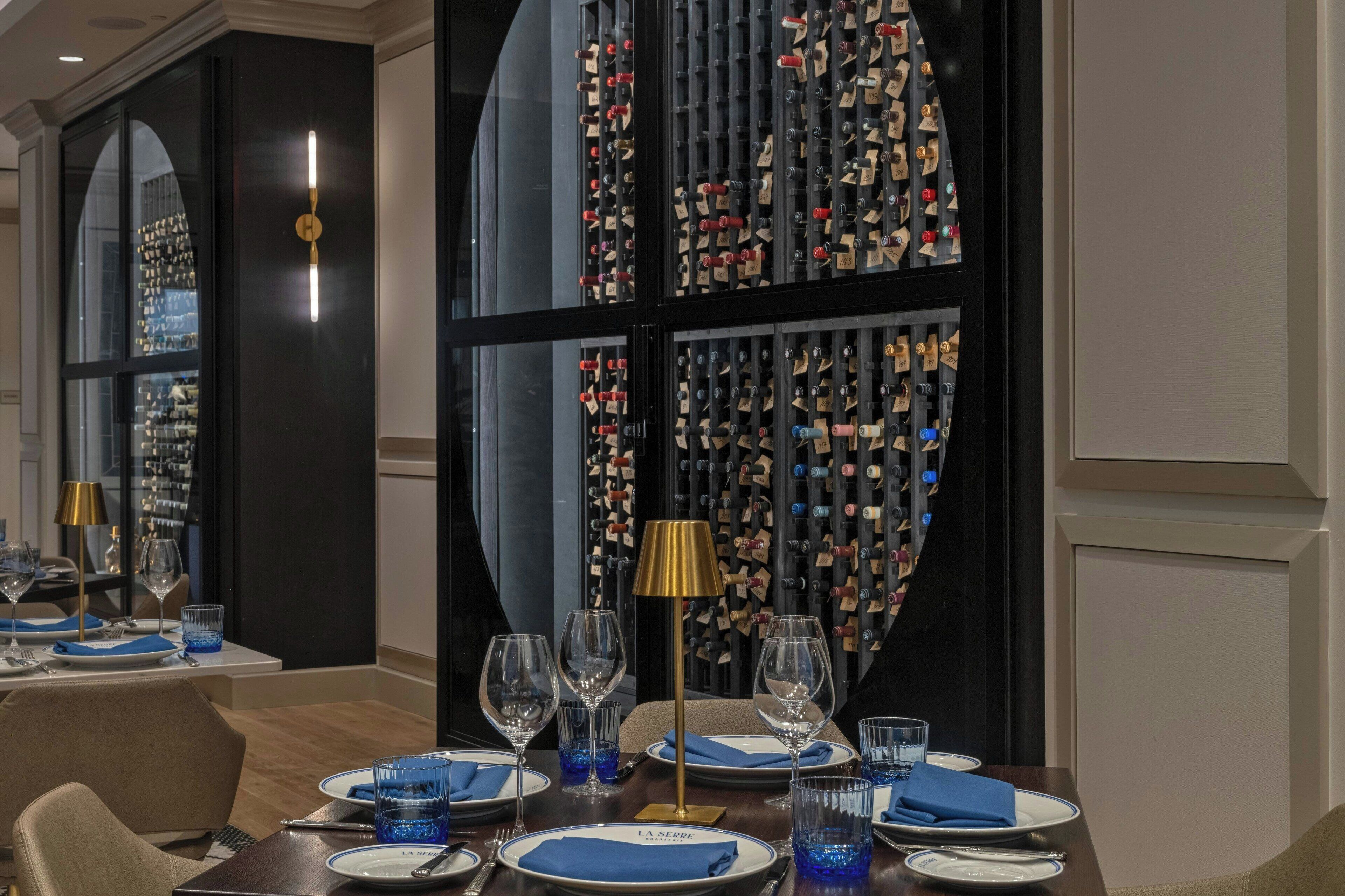 Comedor con una pared de botellas de vino, una lámpara dorada y servilletas azules sobre la mesa.
