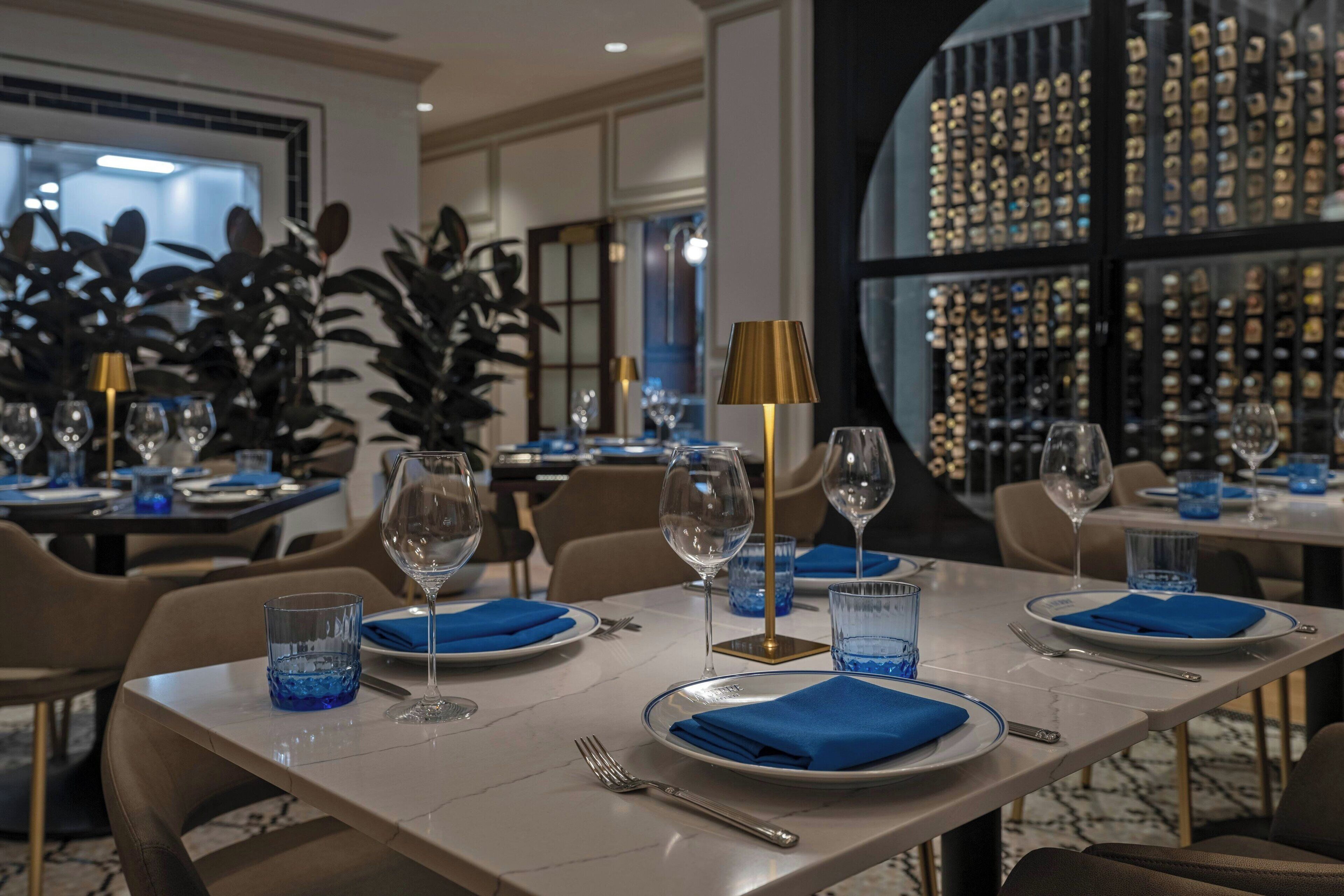 A dining area with tables set for four, each with blue napkins, clear glasses, and a gold-colored lamp.