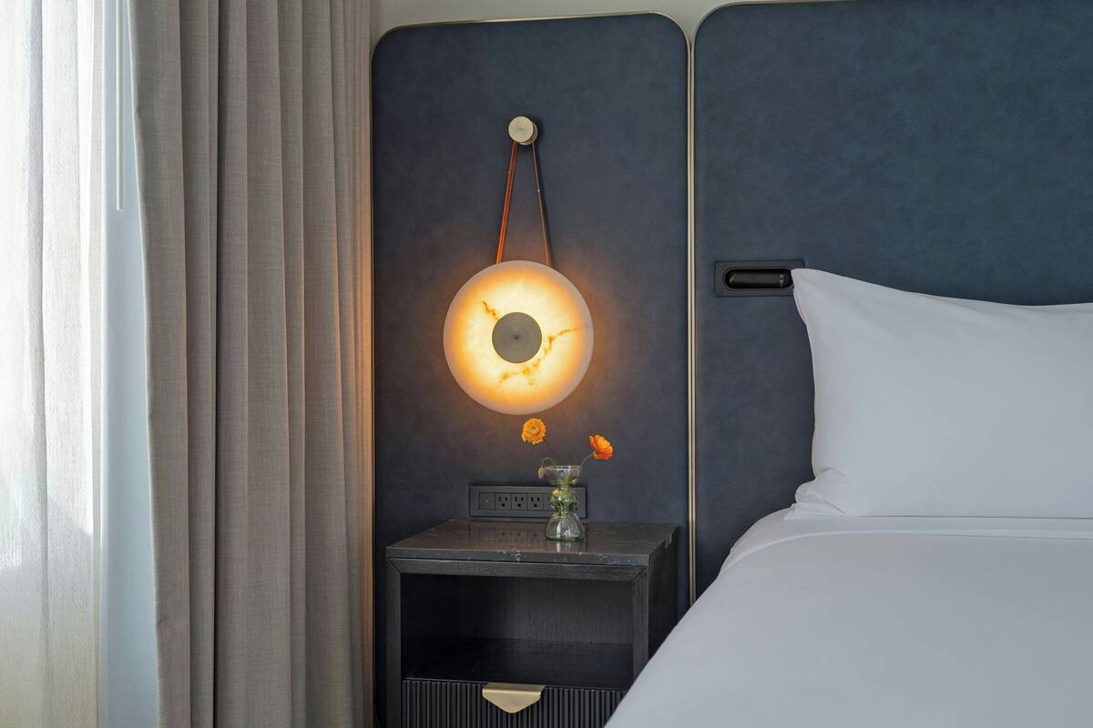 Een opgemaakt bed met een wit kussen en een donker hoofdbord. Een nachtkastje met een lamp en een bloemenvaas is zichtbaar.