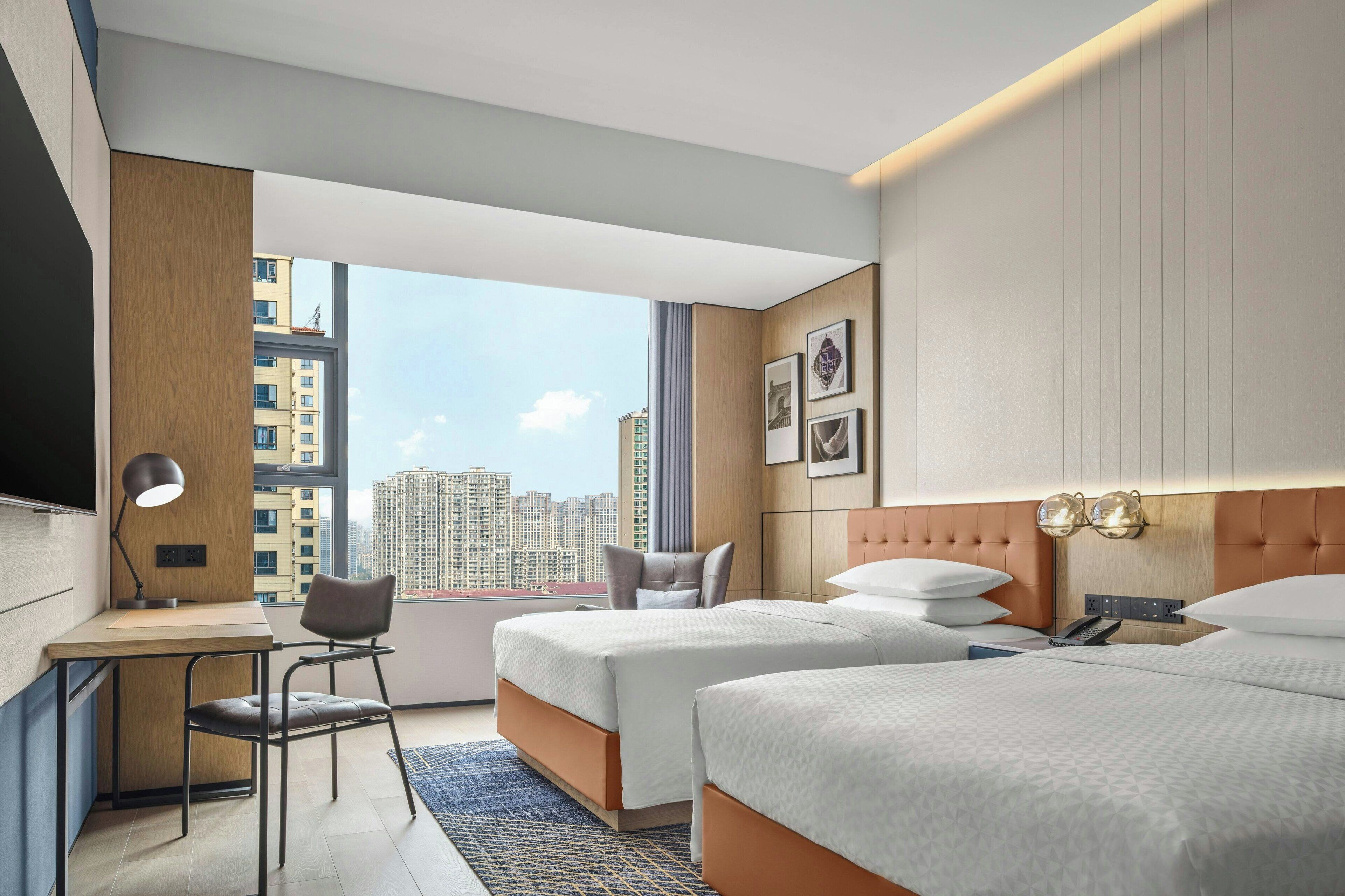 Une chambre d’hôtel avec deux lits, un bureau, une chaise, une télévision et une grande fenêtre donnant sur la ville.