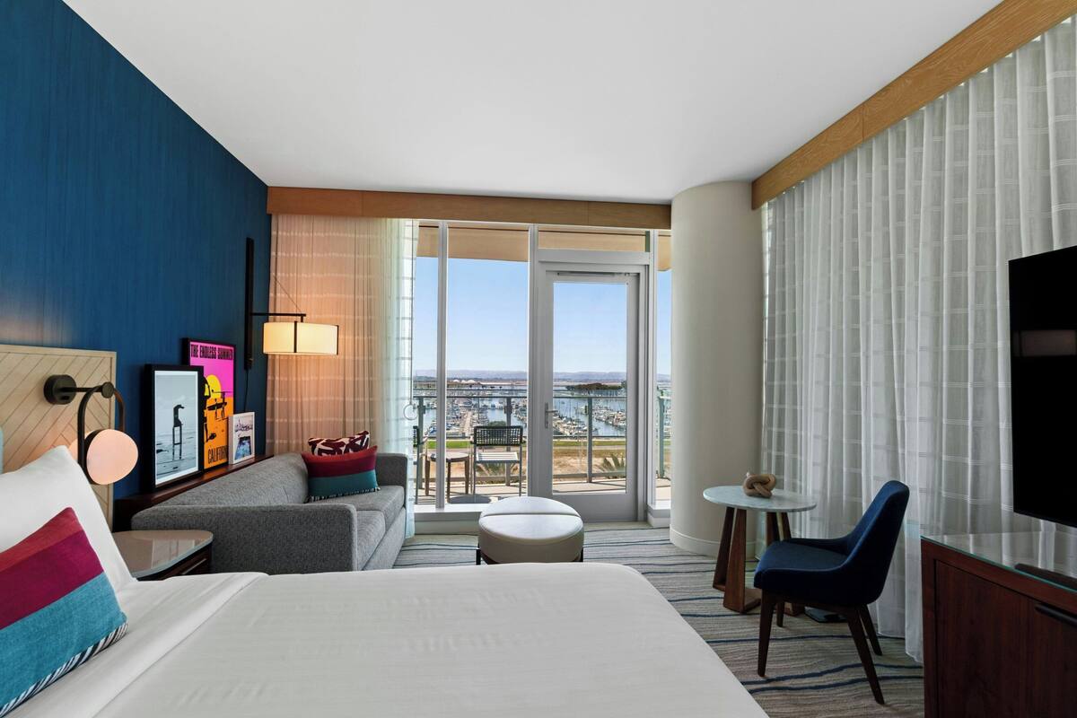 Hotelkamer met een groot bed, een bank, een kleine tafel, een stoel en een televisie. Er is een balkon met uitzicht op de stad.