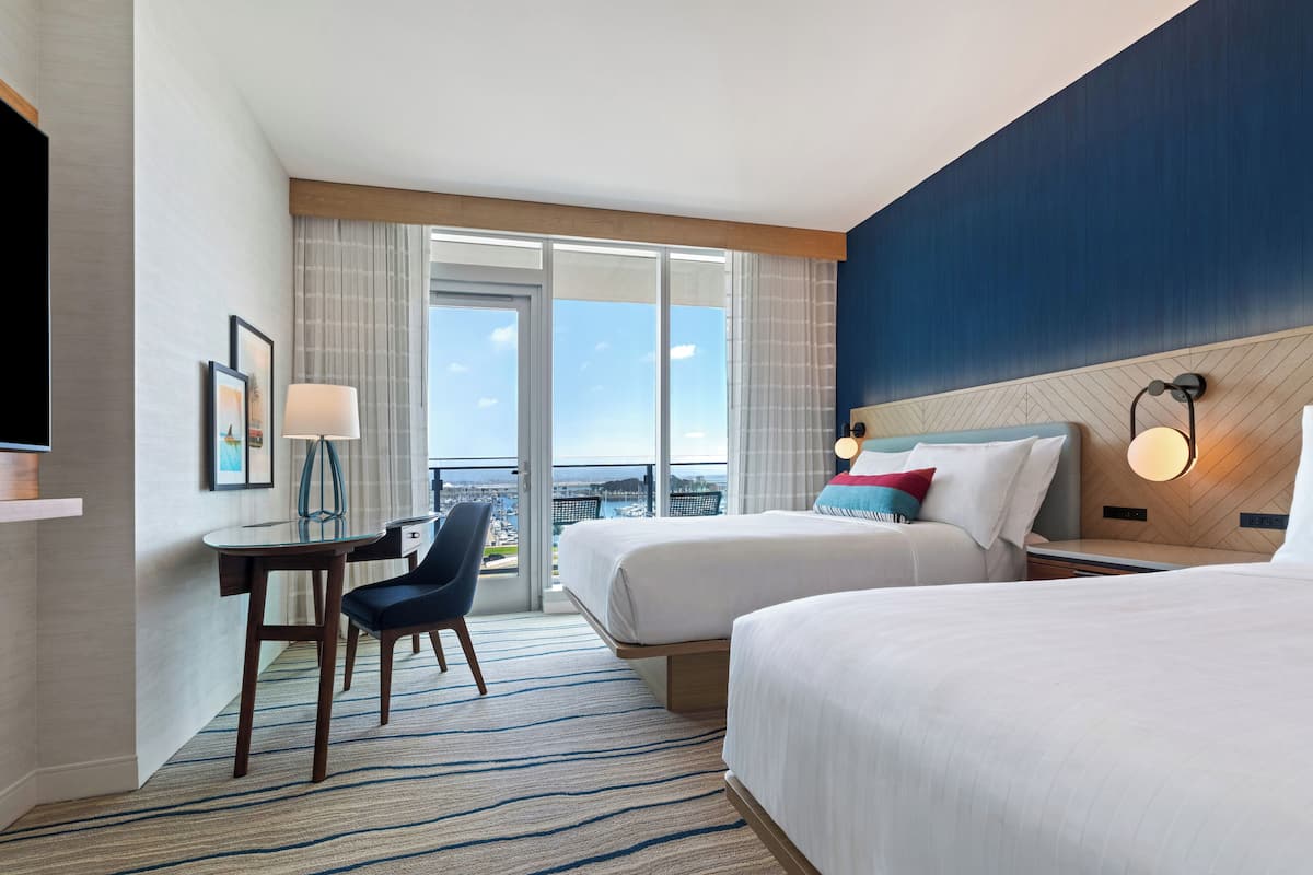 Een hotelkamer met twee bedden, een bureau met stoel, een televisie en een groot raam met uitzicht op de stad.