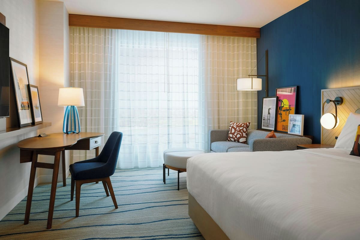 Hotelkamer met een groot bed, een bureau met stoel, een kleine tafel en een bank. Er is een raam met gordijnen, een wandlamp en ingelijste foto's aan de muur.