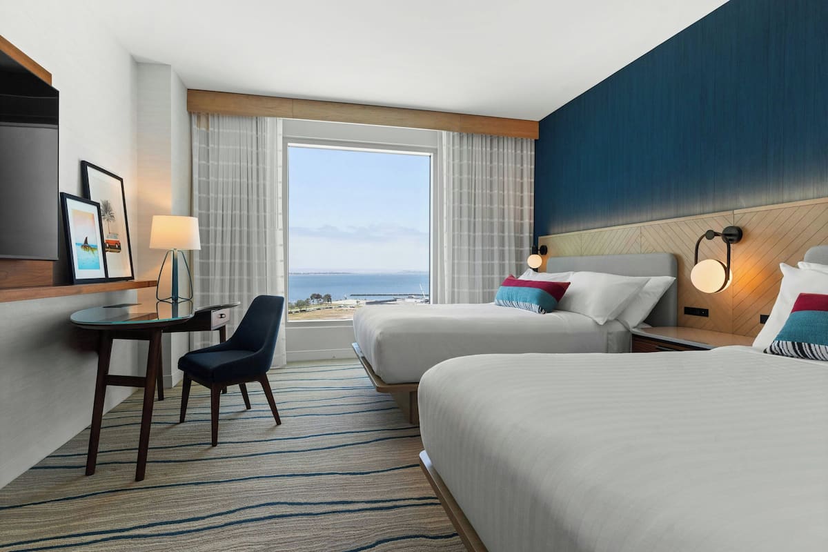 Een hotelkamer met twee bedden, een bureau, een stoel, een televisie en een groot raam met uitzicht op de oceaan.