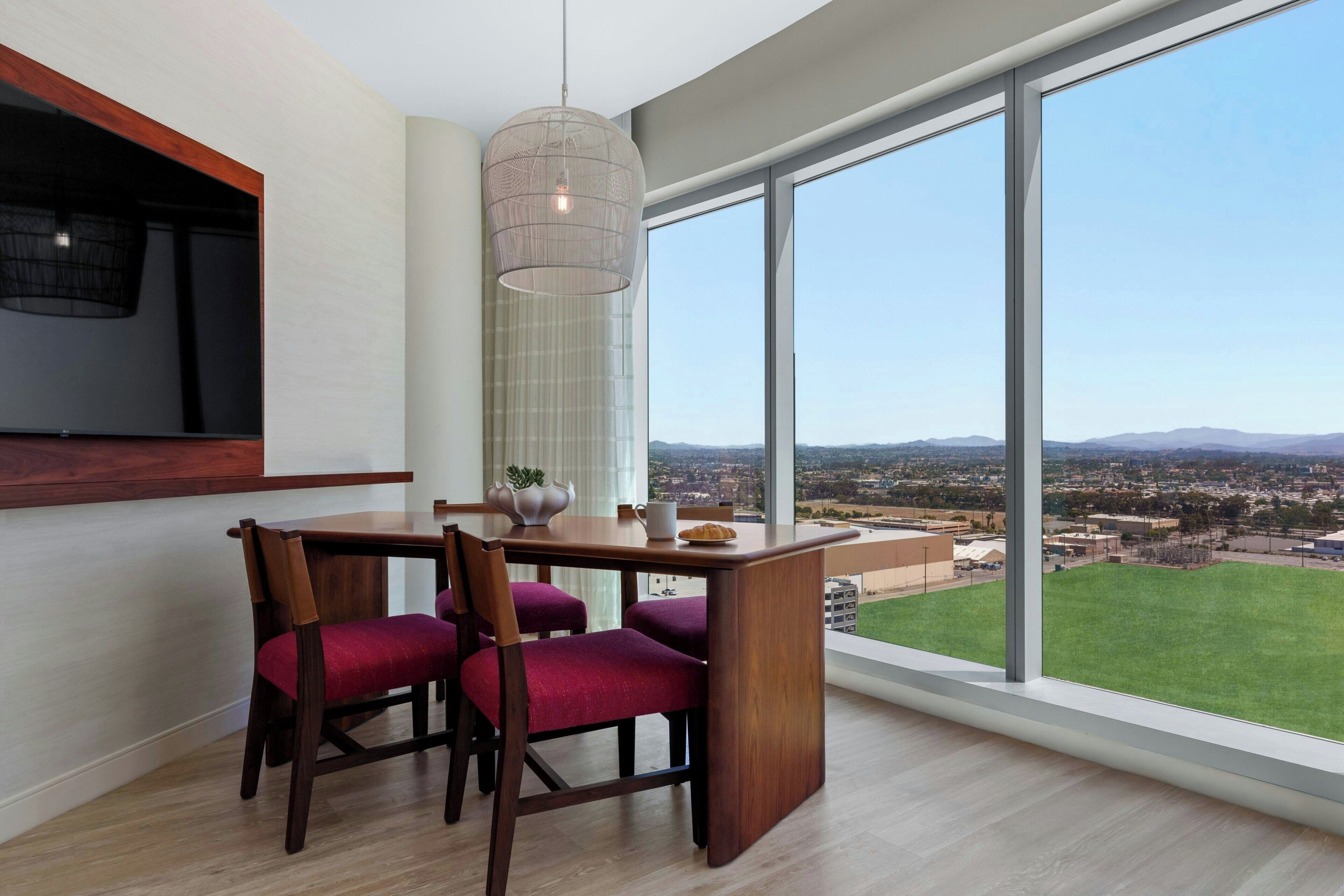 Un área de comedor con una mesa y sillas, una lámpara colgante y un ventanal con vistas a un paisaje urbano y un campo verde.