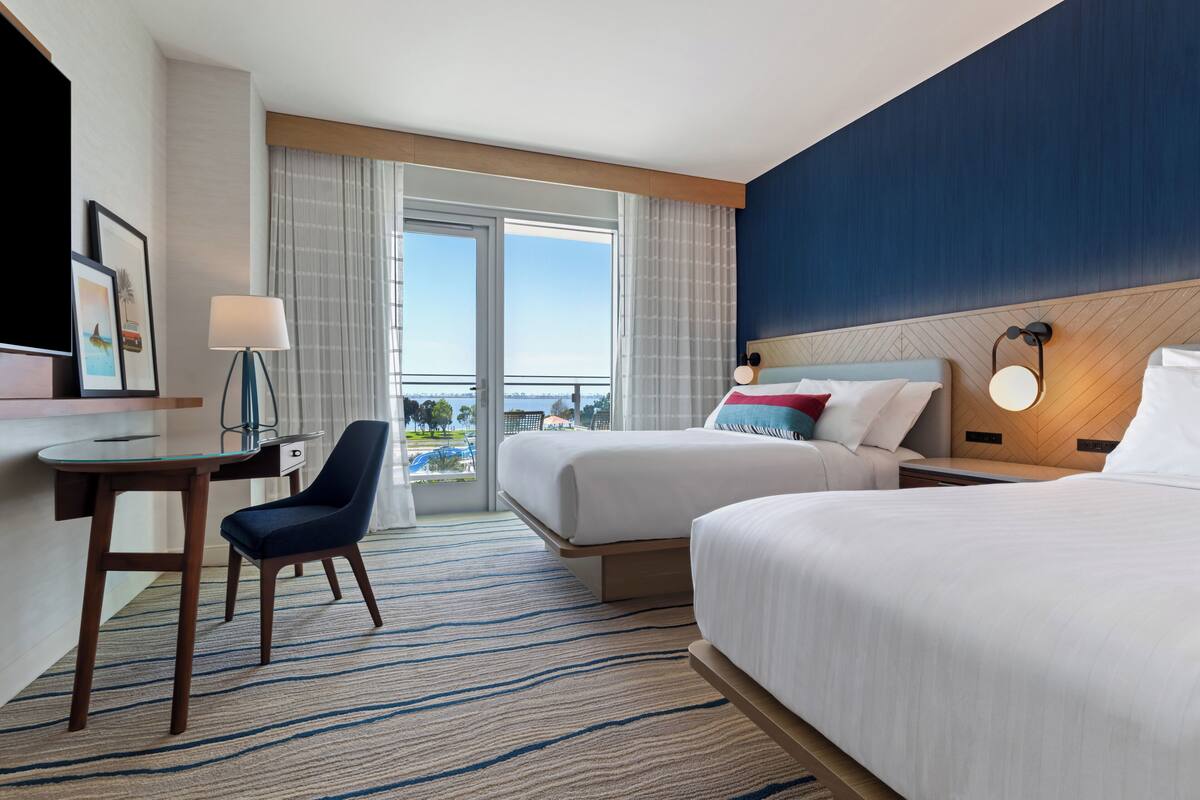 Een hotelkamer met twee bedden, een bureau, een stoel, een televisie en uitzicht op de omgeving.