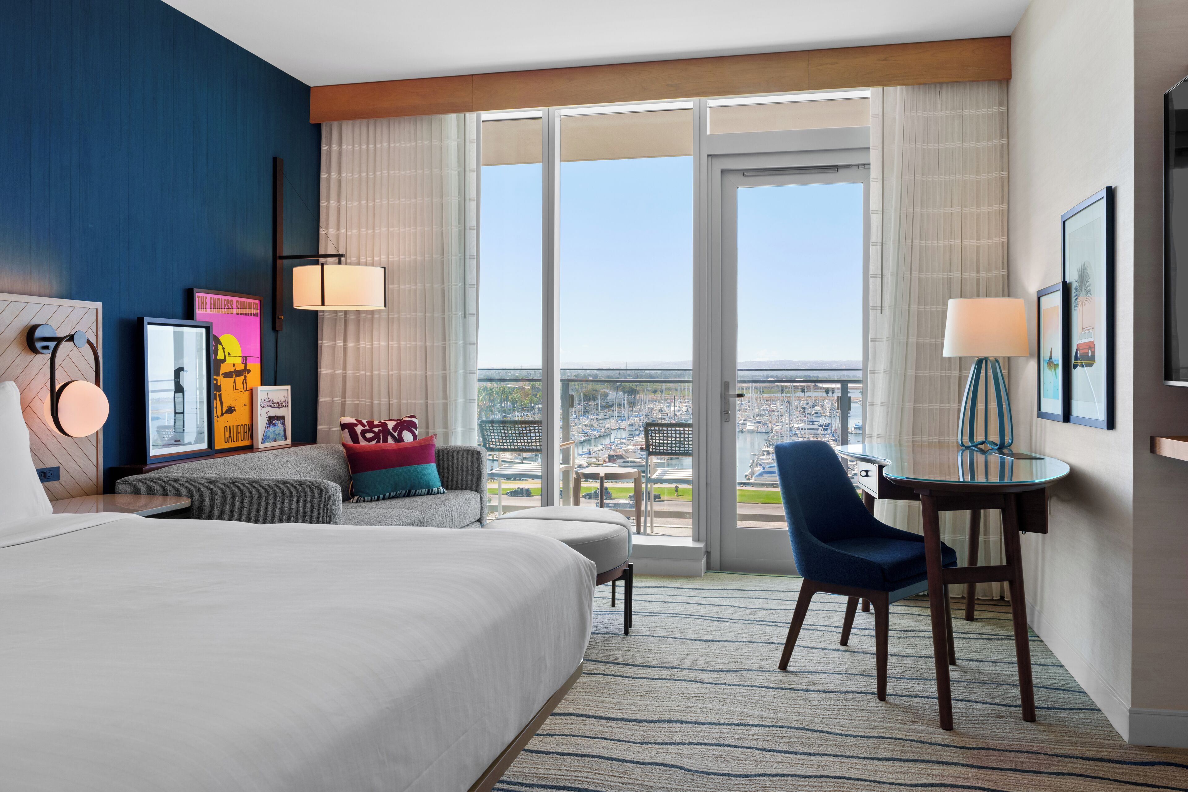 Habitación de hotel con una cama grande, un escritorio con una silla, un sofá y vistas a la ciudad a través de amplios ventanales.